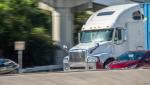 White tractor trailer rig driving under highway underpass with red car on its right.