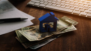 3D house on pile of money with tax and financial forms on the desk.