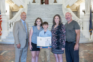 Senator Pat Stefano stands with Luke Hamaker and his family in the capitol rotunda, posing with a certificate from the state senate.
