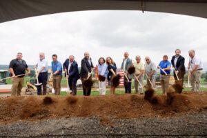 Community leaders and local officials participate in a ceremonial groundbreaking for the Bette Brown Slayton Business Park, standing in a line with shovels as they toss dirt during the event.